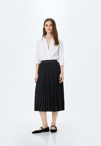 Falda negra plisada con una cinturilla ajustada, combinada con una camisa blanca de botones. La modelo lleva zapatos planos negros con una correa.