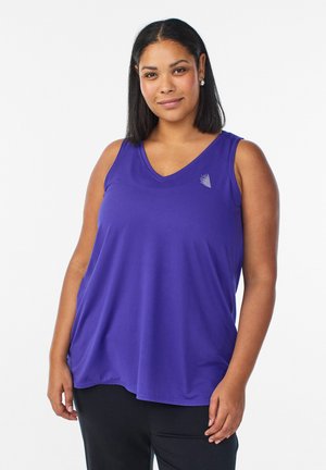 Frau mit glatten schwarzen Haaren trägt ein lila ärmelloses V-Ausschnitt-Oberteil und schwarze Hose, steht vor einem weißen Hintergrund mit entspanntem Ausdruck.