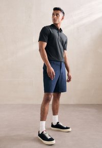 Melnā polo krekls un tumši zila šorti, apvienoti ar tumšiem sporta apaviem, baltām zeķēm. Modelis pozē pie neitrālas sienas dabīgajā apgaismojumā.