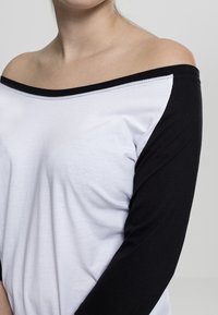 Hvit bomullskjorte med svarte lange ermer, med av-shoulder-design og svart søm langs halslinjen.