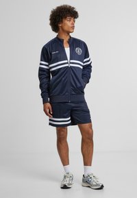 Survêtement bleu marine avec des bandes blanches, composé d'une veste zippée et d'un short assorti. Fabriqué dans un tissu lisse, avec un logo sur la poitrine.