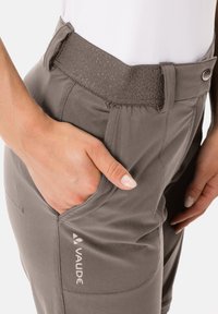 Grau, leichte Outdoor-Hosen mit einem strukturierten elastischen Bund, seitlichen Taschen und VAUDE-Logo. Nahaufnahme einer Hand, die auf der Tasche ruht.