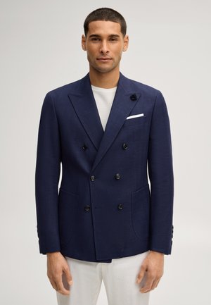 Man draagt een navy dubbelgebreid colbert met een wit pochet en witte broek, staand voor een effen lichte achtergrond.