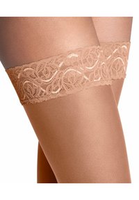 Bas autofixants beige montant jusqu'à la cuisse, avec un haut en dentelle décorative présentant des motifs et volutes complexes, affichant une texture lisse et mate.