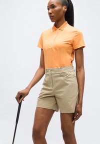 Γυναίκα που φορά πορτοκαλί πόλο μπλούζα και μπεζ σορτς, κρατώντας ένα γκολφ κλαμπ, στέκεται μπροστά σε ένα απλό φωτεινό φόντο.