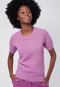 Vaaleanlila, lyhythihainen neulepusero, jossa on timanttikuvio ja ribatty pääntie sekä helma. Rennon istuvuuden omaava, käytetty kädet taskuissa.