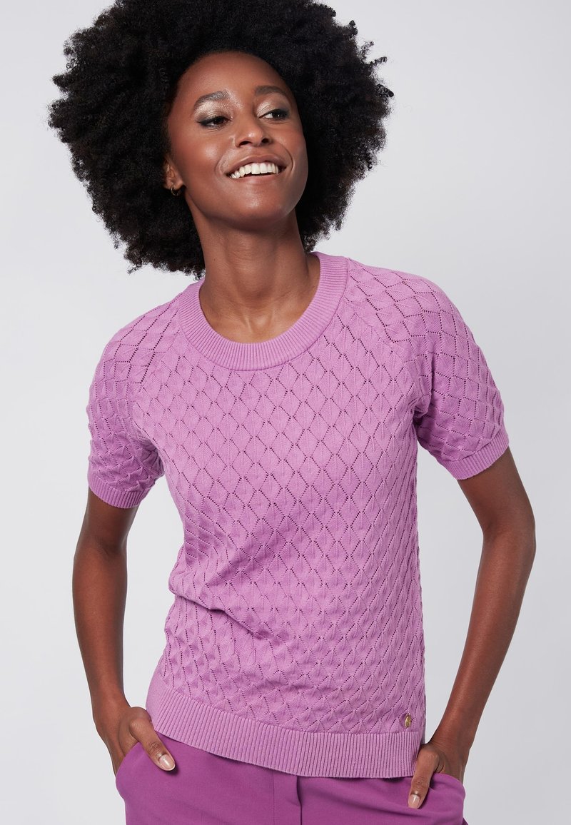 Vaaleanlila, lyhythihainen neulepusero, jossa on timanttikuvio ja ribatty pääntie sekä helma. Rennon istuvuuden omaava, käytetty kädet taskuissa.