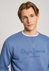 Sudadera azul claro con un acabado texturizado, que presenta un logotipo bordado en azul oscuro "Pepe Jeans LONDON" en la parte frontal. Cuello redondo y puños de canalé.