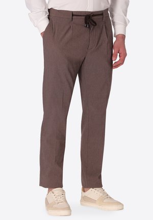 Uomo che indossa pantaloni marroni strutturati con coulisse nera e sneaker beige, in piedi con una mano in tasca.