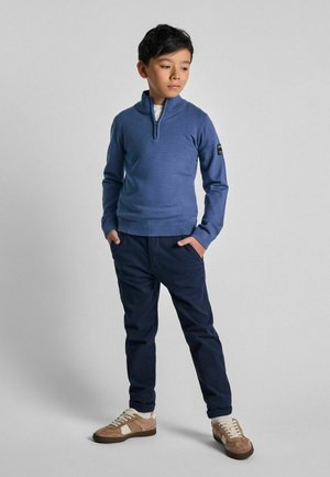 Pull à col zip bleu clair, pantalon marine et baskets beiges avec des accents blancs. Le pull présente une coupe ajustée et des poignets côtelés.