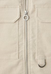 Tela de pana beige con cremallera metálica central con tirador circular y dos bolsillos delanteros, uno con detalle de anilla metálica en forma de D.