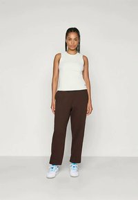 Hvit ermeløs topp og brune bukser, med en glatt tekstur. Hvite sneakers med blå detaljer fullfører antrekket. Modellen står rett.