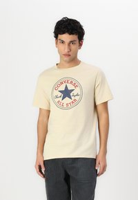 Camiseta de algodón beige con un gráfico redondo que presenta una estrella azul y el texto "Converse All Star" en rojo y marino. Mangas cortas, cuello redondo.