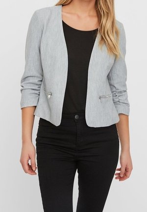 Kobieta w dopasowanej, jasnoszarej marynarce z zamkami błyskawicznymi, na czarnej bluzce i czarnych spodniach, stoi z ramionami luźno opuszczonymi wzdłuż ciała.