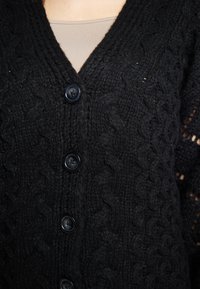 Cardigan nero lavorato a maglia con scollo a V, motivo testurizzato e cinque grandi bottoni neri sulla parte frontale. Dettagli di lavorazione aperta sulle maniche.