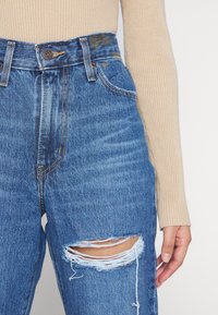 Jeans de mezclilla azul de cintura alta con un diseño desgastado que presenta una gran área rasgada en la rodilla izquierda, combinados con una blusa beige de canalé.
