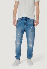 Jeans de mezclilla azul claro rasgados con un ajuste relajado, que cuentan con detalles deshilachados y parches decorativos, combinados con una camiseta blanca de manga corta y zapatillas blancas.