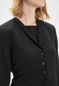 Blazer noir ajusté avec un col cranté, des épaulettes et quatre boutons, présentant une texture lisse et une silhouette cintrée.