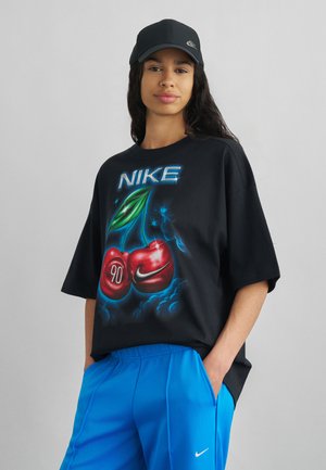 Kvinde iført sort Nike-kasket, sort t-shirt med stort kirsebærmotiv og teksten "NIKE", kombineret med lyseblå Nike-bukser, hænderne i lommerne.