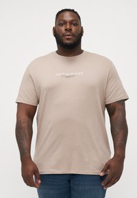 Bež majica kratkih rukava s okruglim izrezom, s natpisom "JACK & JONES Originals" otisnutim bijelom bojom na prednjoj strani. Mekani pamučni materijal.