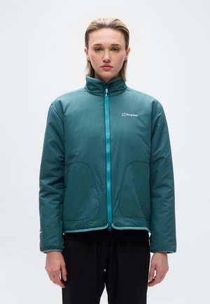 Jaunas žmogus vilki žalią Berghaus striukę su užtrauktuku ir juodas kelnes, stovi priešais šviesų ir paprastą foną, žiūri tiesiai, veide neutrali išraiška.