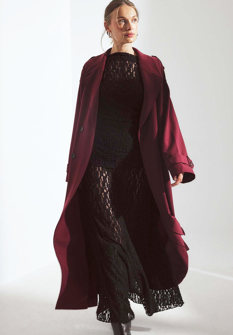 Manteau long bourgogne avec de grands revers et une robe en dentelle noire en dessous. La dentelle a un motif texturé avec des manches longues et une coupe fluide.