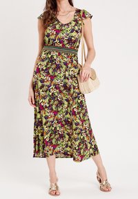 Maxikleid mit floralem Muster in Grün-, Lila- und Gelbtönen; Rüschenschultern; tailliert; kombiniert mit einer beigen, gewebten Handtasche und Sandalen.