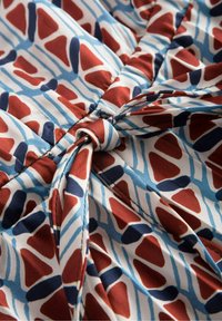 Tištěná látka má vícebarevný geometrický vzor v červené, modré a bílé barvě, s uzlovým detailem uprostřed. Měkká textura.