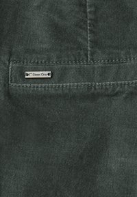 Mörkgrön texturerad tyg med en metallisk "Street One"-etikett sydd på bakfickan, med synliga sömdetaljer.