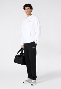 Weißer Kapuzenpullover mit Reißverschluss und Kordeln, schwarze Jogginghose mit Logo und graue Sportschuhe. Hält eine schwarze Reisetasche. Glatte Texturen.