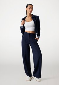 Marineblauwe trainingsjack met witte strepen, gecombineerd met hoge taille wijde pijpen. Witte crop top zichtbaar. Glad materiaal, minimale branding.