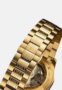 Gouden metalen horloge met een patroonarmband. Heeft geometrische gravures en een transparante achterkap die ingewikkelde mechanica onthult.