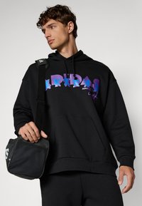 Sweatshirt preta com texto gráfico em roxo e azul; possui um bolso tipo canguru e um corte relaxado. Os acessórios incluem uma bolsa de ombro preta.