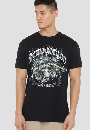Black cotton t-shirt featuring a graphic print of a creature with a flower head, text "Demogorgon" and tour details in white and colorful accents.