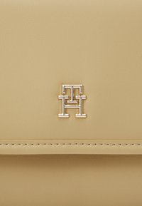 Surface en cuir beige lisse avec un petit logo "TH" en métal doré entrelacé et une couture horizontale sous le logo.