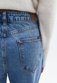 Blå denimjeans med hög midja, bakfickor och bruna läderdetaljer vid midjan. Sömnadsdetaljer i kontrasterande tråd.