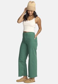 Jeans verde a gamba larga con orli sfrangiati, abbinati a una canotta bianca e a un berretto in maglia beige. Le scarpe slip-on color beige completano il look.