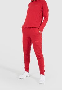 Ensemble sweat à capuche rouge et pantalon de jogging en tissu doux. La capuche dispose d'une poche kangourou et d'un logo, tandis que les joggers ont des poches latérales et des jambes fuselées.