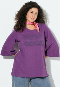 Camiseta de algodón púrpura con mangas acampanadas anchas, con el texto "LOOK GOOD" en estampado oscuro. Combinada con una bufanda rosa con estampado.