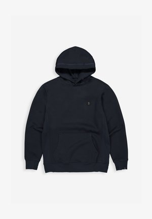 Man draagt een marineblauwe hoodie en bijpassende broek, staat met ontspannen handen, licht verband om linkerhand, neutrale grijze achtergrond.