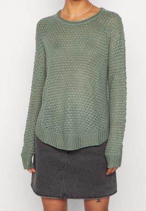 Personne portant un pull en tricot vert olive texturé et une jupe en jean gris foncé, avec un petit tatouage visible sur la cuisse gauche.