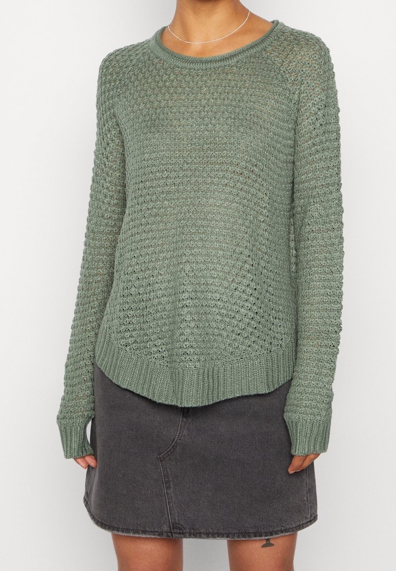Personne portant un pull en tricot vert olive texturé et une jupe en jean gris foncé, avec un petit tatouage visible sur la cuisse gauche.