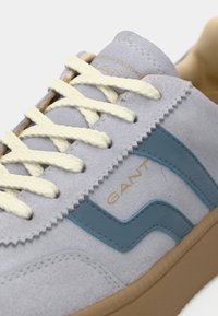 Αθλητικό παπούτσι από ανοιχτό γκρι σουέτ με μπλε δερμάτινες λεπτομέρειες, κρεμ κορδόνια και καφέ καουτσούκ σόλα. Διαθέτει ανάγλυφο λογότυπο της μάρκας στο πλάι.