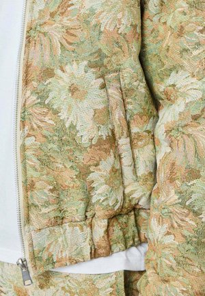 Gros plan sur une manche de veste à motifs floraux avec un poignet côtelé dans des tons beiges, verts et blancs, à côté d'une fermeture éclair blanche partiellement visible.