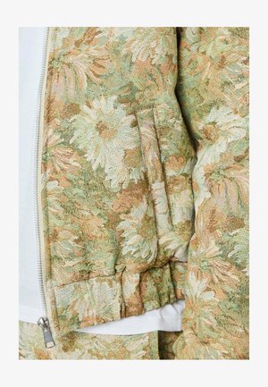 Gros plan sur une manche de veste à motifs floraux avec un poignet côtelé dans des tons beiges, verts et blancs, à côté d'une fermeture éclair blanche partiellement visible.