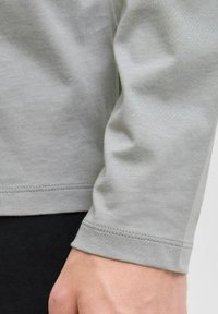 Close-up van een lichtgrijze lange mouw shirt. Zachte, gladde katoenstof met zichtbare stiksels. Naaddetails en lichte textuurvariaties zijn aanwezig.