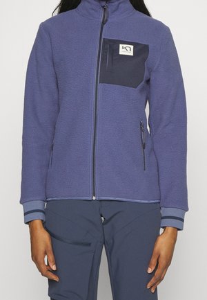 Osoba ubrana w niebieską kurtkę polarową z czarną kieszenią na piersi i zamkiem błyskawicznym, połączona z ciemnoniebieskimi spodniami z boczną kieszenią na zamek.