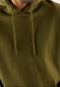 Felpa verde oliva con una texture morbida, dotata di tasca frontale, cordini regolabili e occhielli in metallo. Design semplice senza decorazioni.