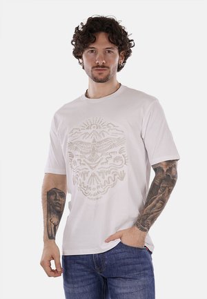 Camiseta de algodón blanca con un diseño gráfico en un beige claro que presenta montañas, un águila y patrones abstractos; mangas cortas y cuello redondo.