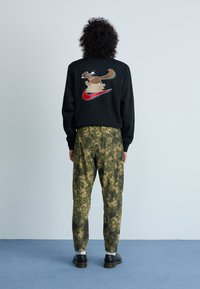 Μαύρη μπομπερ sweatshirt με γραφικό κινούμενων σχεδίων και λογότυπο στην πλάτη, σε συνδυασμό με παντελόνι σε παραλλαγή πράσινου. Μαύρα παπούτσια ολοκληρώνουν το σύνολο.